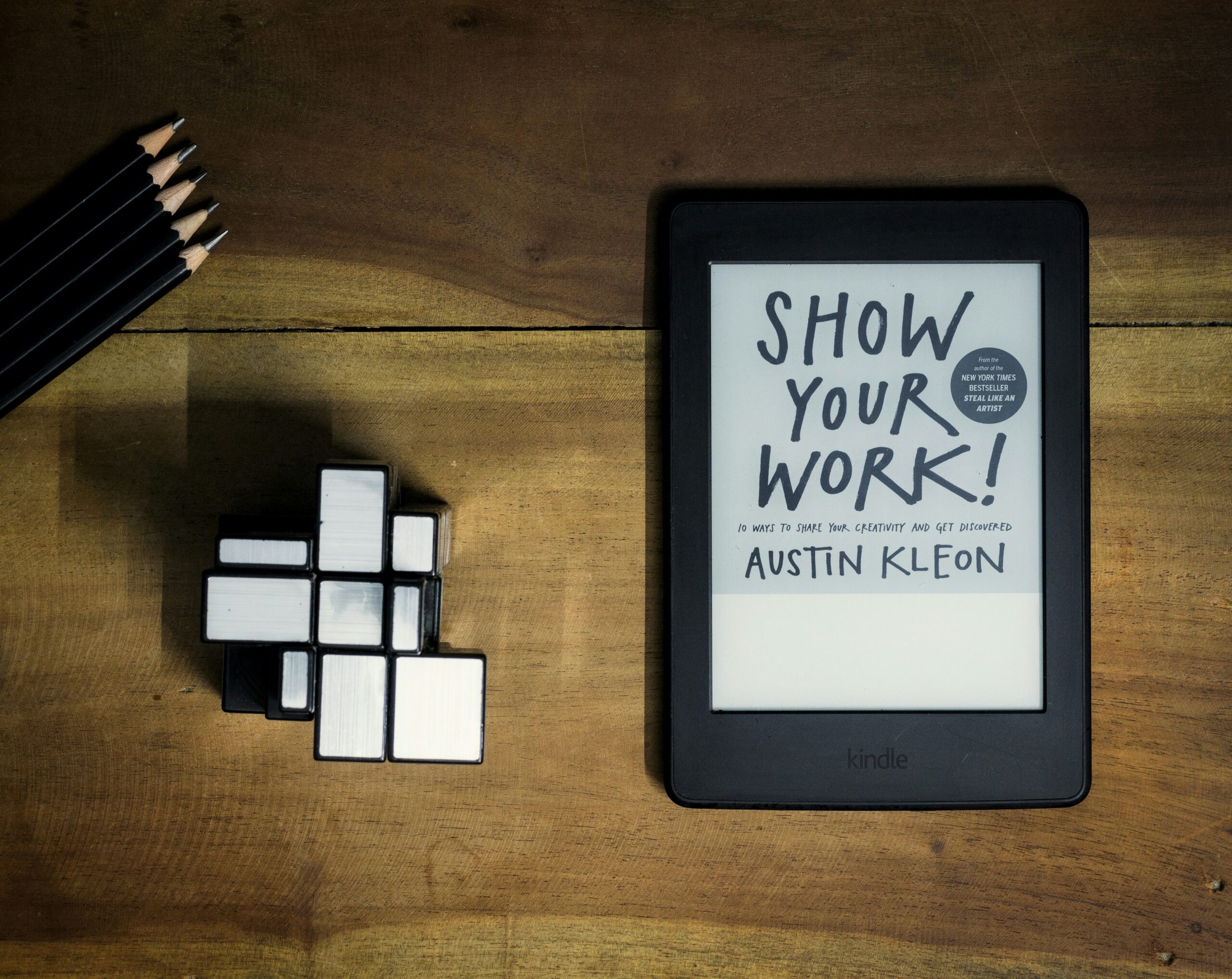 Kindle on a wooden desk with an ebook open. A Mirror- Rubik's cube and some pencils are also on the desk.